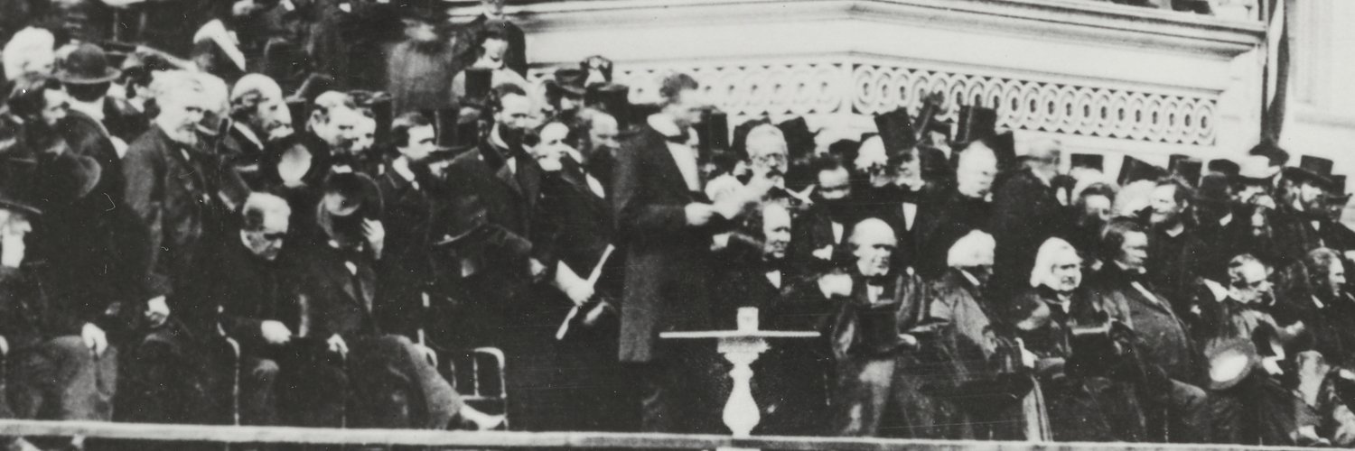 Abraham Lincoln, bareheaded, reads from a paper at a small table on the crowded east portico of the Capitol, among seated officials in top hats