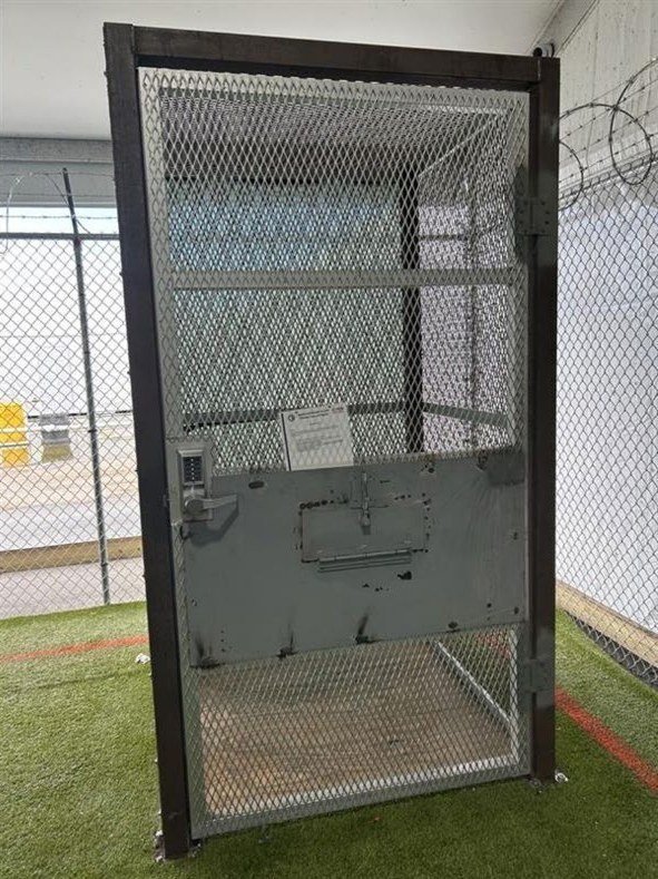 A narrow metal-mesh cage with a solid door, standing on artificial turf inside a fenced enclosure