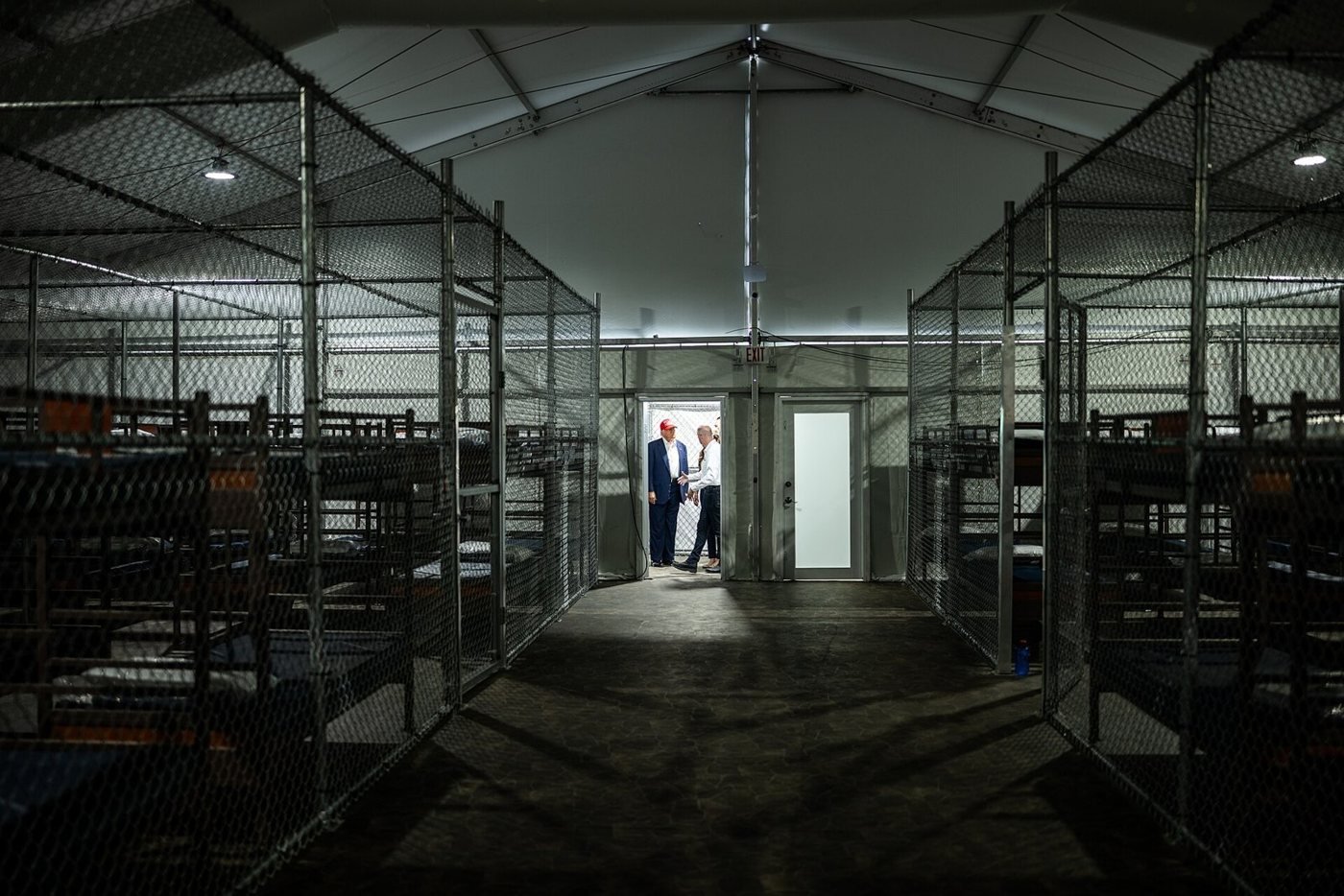 Rows of empty bunk beds inside wire-mesh cages under a white tent roof, with figures standing in a lit doorway at the far end