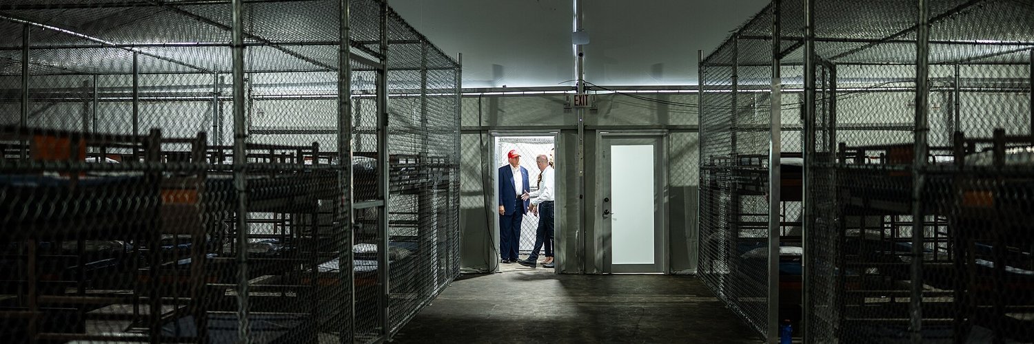 Two rows of chain-link cages holding bunk beds inside a tent, with the President and an official standing in a lit doorway at the far end
