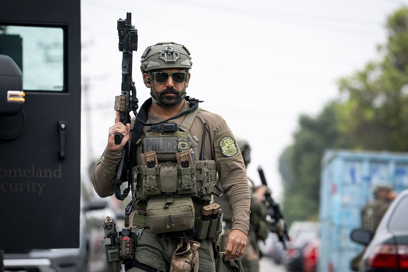 A bearded federal agent in a helmet, sunglasses and body armour marked POLICE holds a rifle upright beside an armoured vehicle