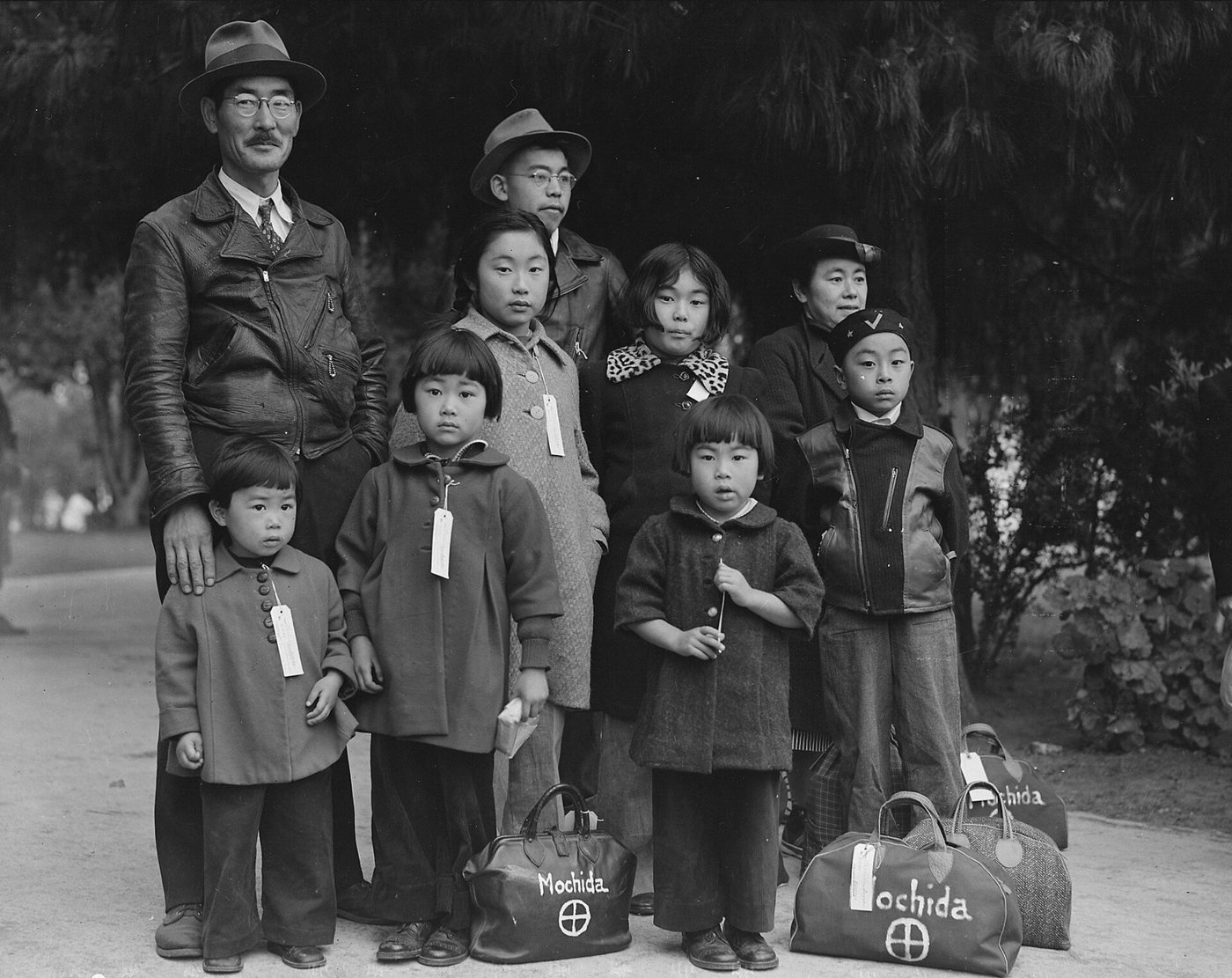 A Japanese American father, mother and children in coats, each wearing an identification tag, stand beside bundled luggage