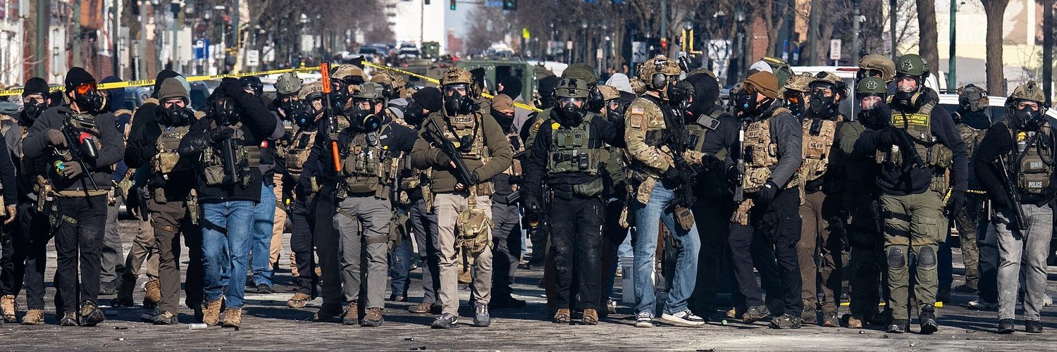 A line of some thirty federal agents in gas masks, helmets and body armour, many holding rifles, stretches across a city street behind police tape