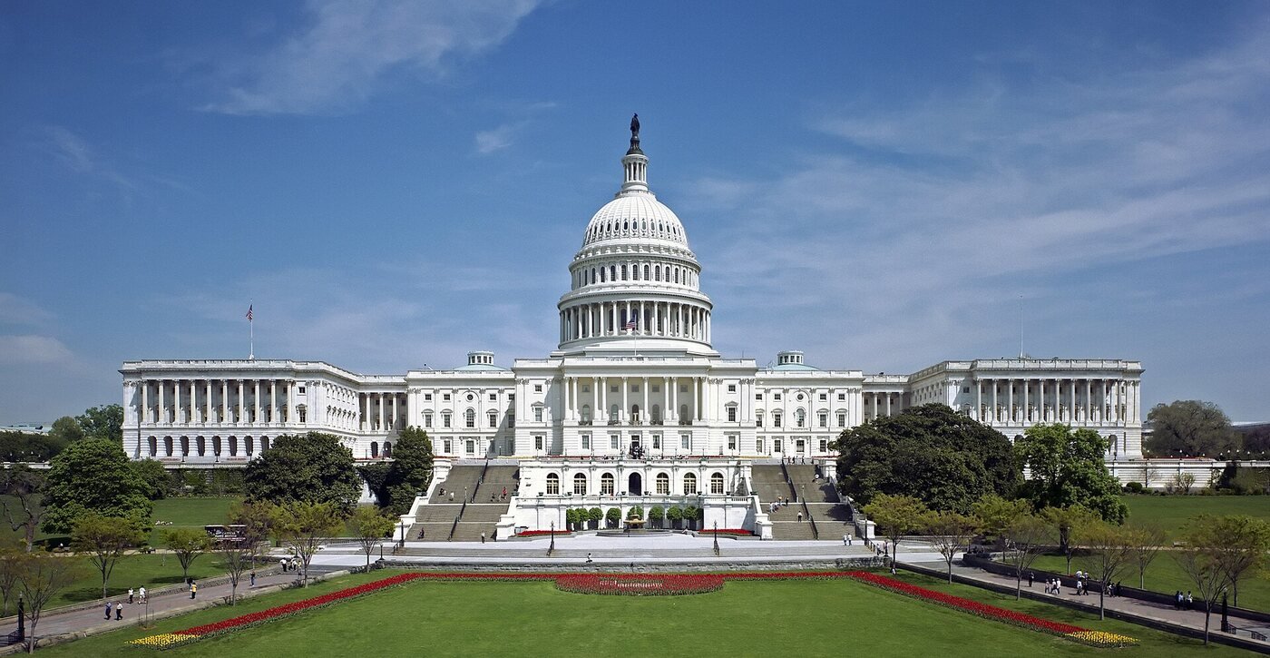 The white marble West Front of the United States Capitol under a blue sky, its dome rising above the terraces and the broad central steps