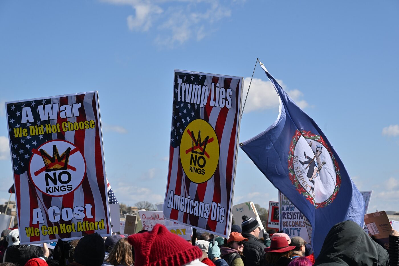 Marchers carry No Kings signs and flags across a bridge under a blue sky