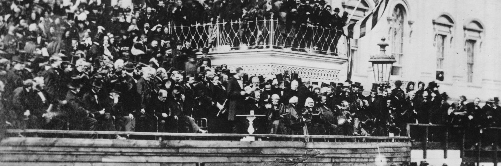 The crowded east portico of the Capitol on 4 March 1865, with Abraham Lincoln standing at a small table reading his second inaugural address, a flag draped from the balcony above