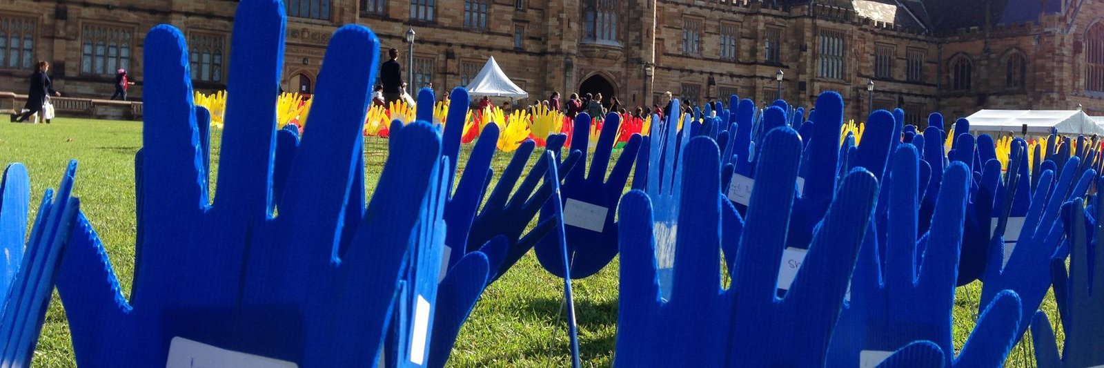 Rows of blue cut-out hands planted on the Quadrangle lawns of the University of Sydney, with red and yellow hands beyond and the sandstone Main Quadrangle behind