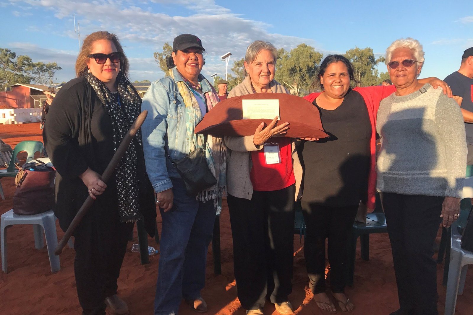 Megan Davis, June Oscar, Pat Anderson holding the Uluru Statement from the Heart in its coolamon, Sally Scales, and Irene Davey standing on red earth at Uluru under a blue sky