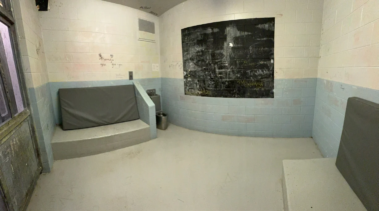 A concrete-block cell with a vinyl mattress on a concrete bench, a second mattress at right, a steel toilet, and a black graffiti-covered panel on the wall