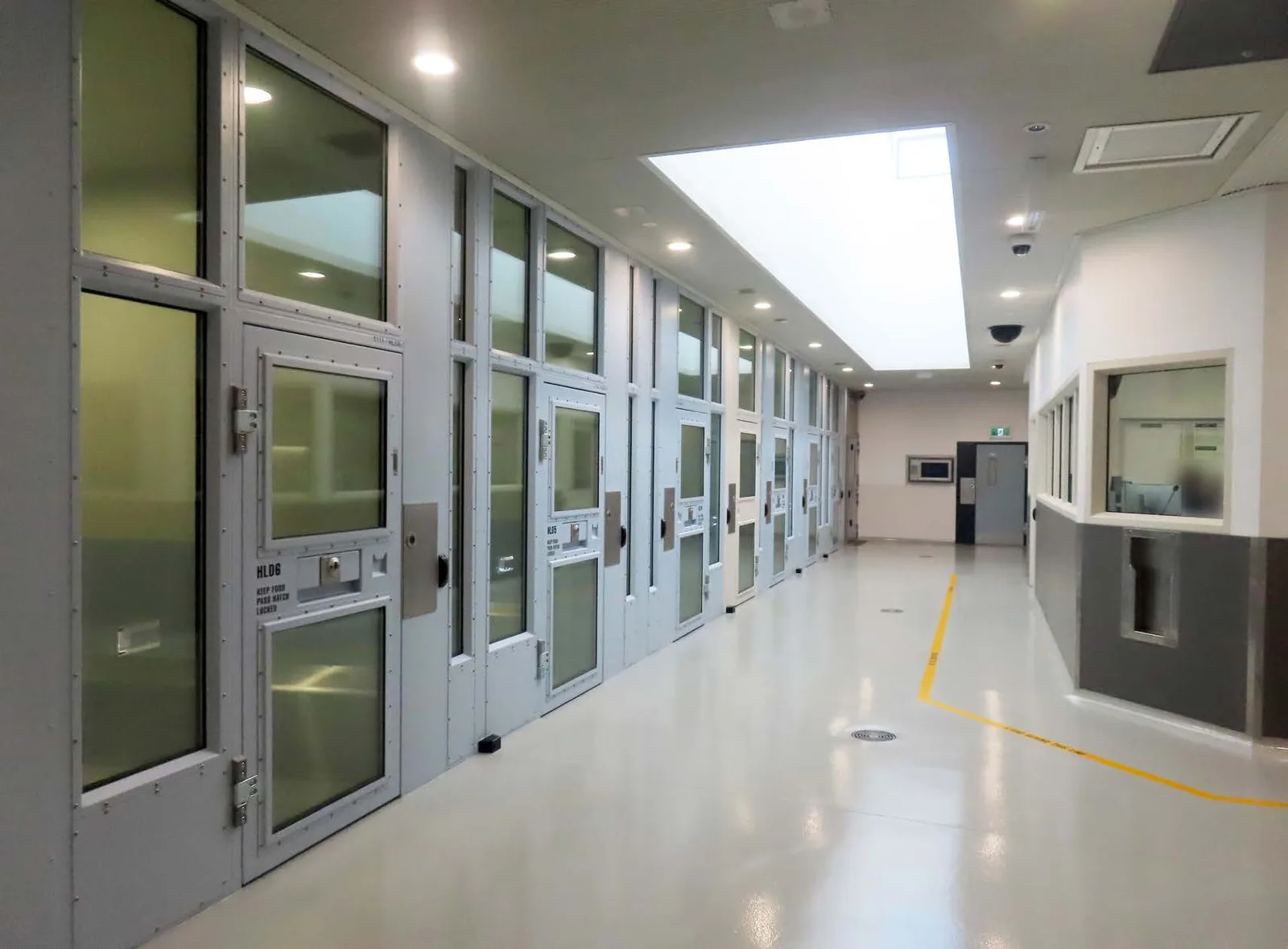 A long, brightly lit corridor of steel-framed holding cell doors with glass panels, a grey floor, and a staff window at the far end