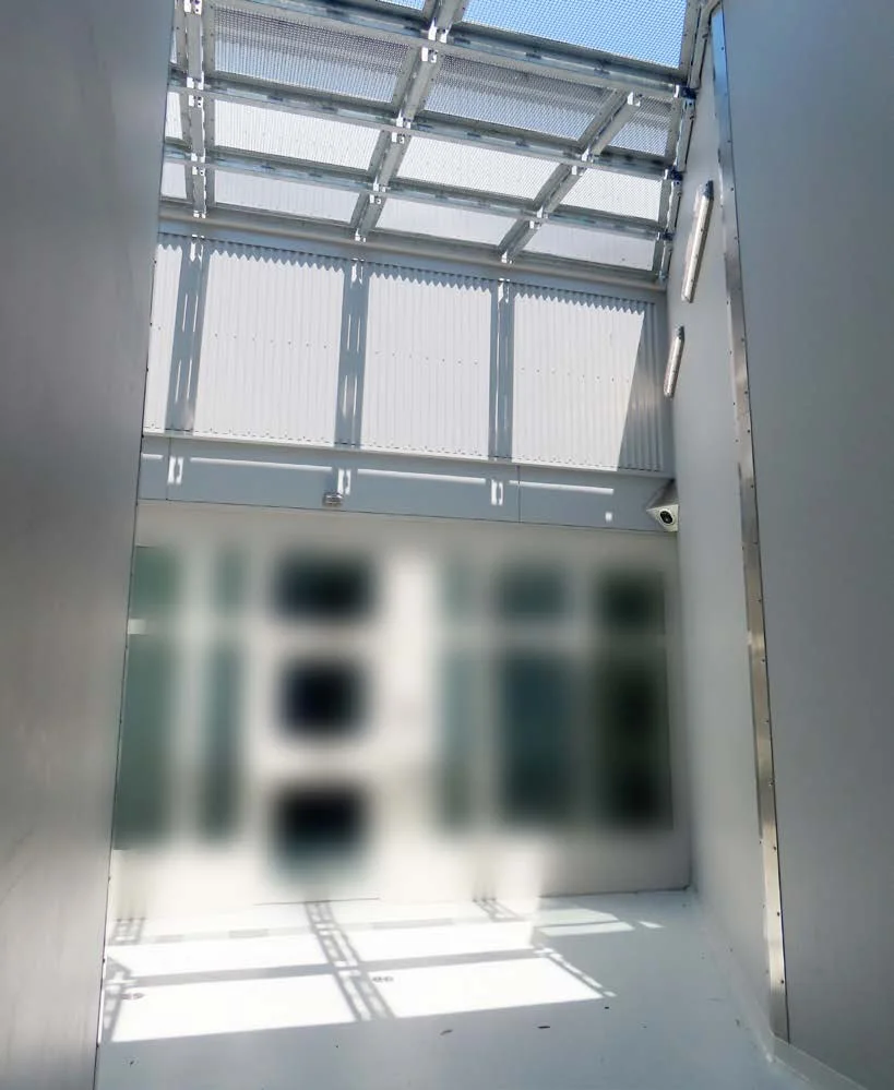 A narrow exercise yard enclosed by high grey walls, with a steel mesh roof above and part of the image blurred